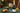 A bottle of Tanners Champagne Extra Reserve stands on a green plaid tablecloth beside two filled champagne flutes, a gold candle holder, a plate of assorted chocolates, and a wrapped gift box, with a decorated Christmas tree in the background.