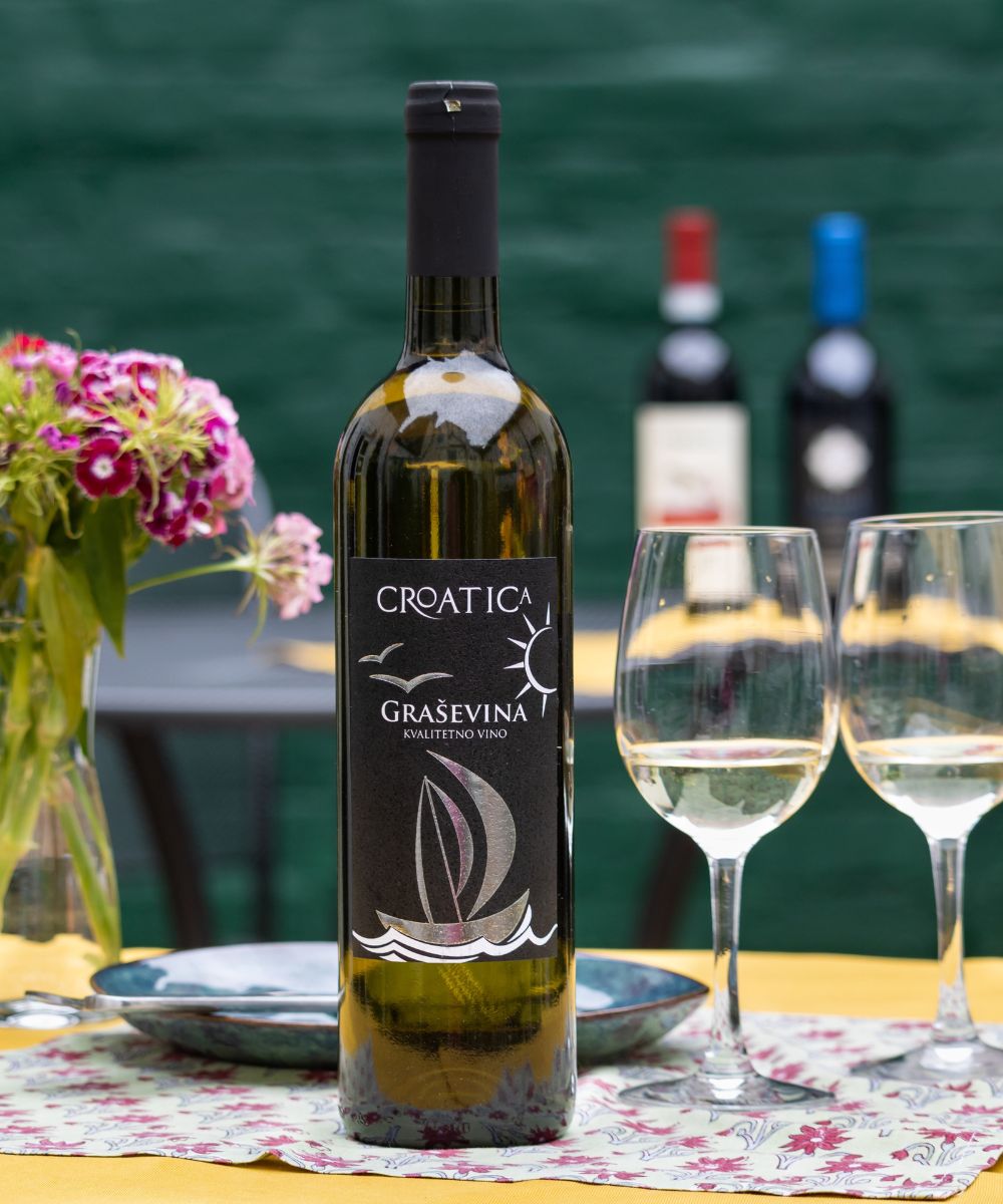 A bottle of Croatica Grasevina, alongside two poured glasses displayed on a table in an outdoor courtyard.