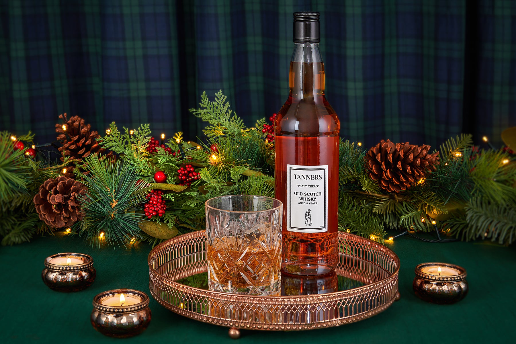 A bottle of Tanners 8 year old Single Malt Scotch whisky stands on a round copper tray next to a cut glass, surrounded by festive Christmas greenery, pinecones, candles, and a dark green plaid backdrop.