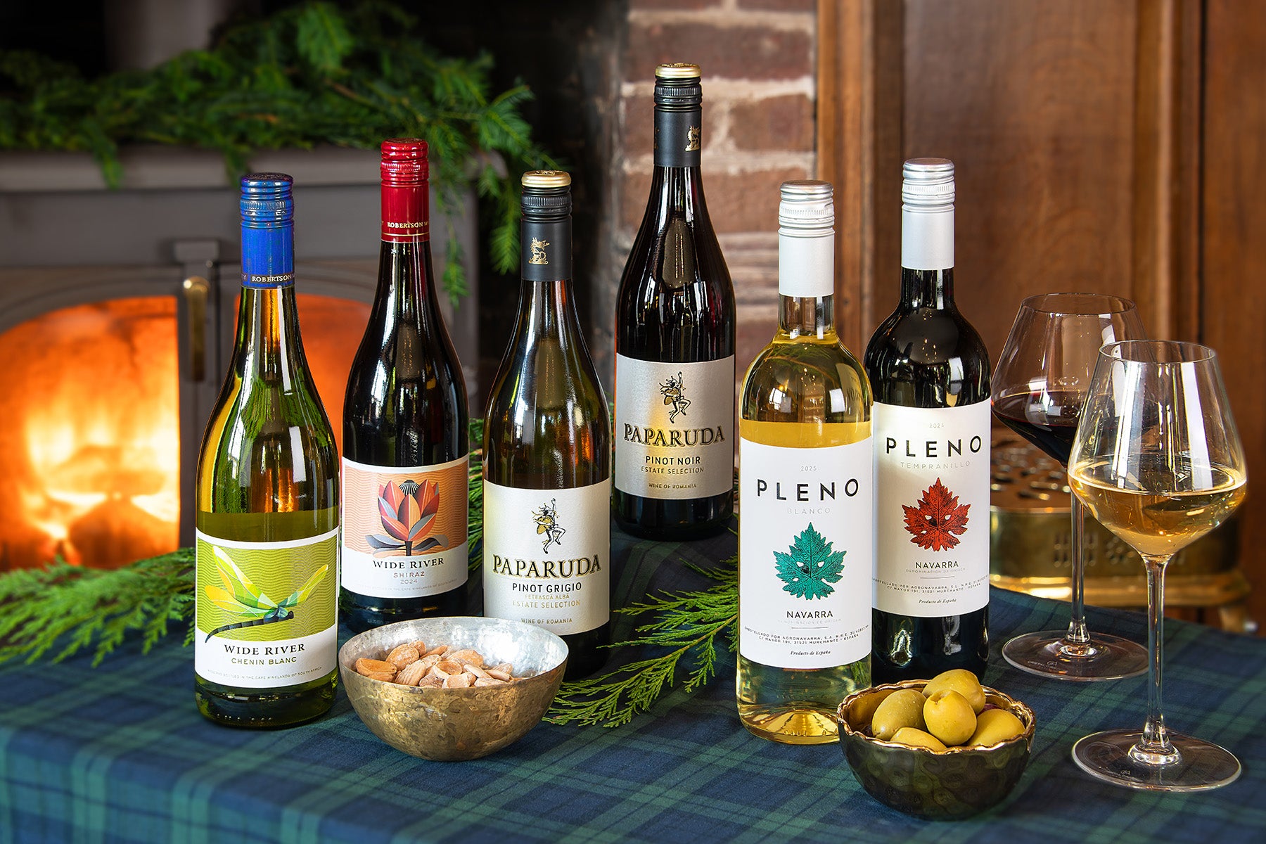 Six wine bottles and two filled wine glasses arranged on a plaid tablecloth with bowls of olives and pine branches in front of a warm fireplace. Included are bottles from Pleno, Parruda and Wide River.